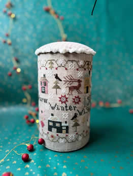 May include: A cylindrical needle minder with a white fabric cover and a pink velvet top. The fabric cover is embroidered with a winter scene including houses, trees, birds, a deer, and the text 'Farm Winter Wishes'. The needle minder is sitting on a teal surface with a gold pattern.